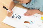 Load image into Gallery viewer, Inside lift‑the‑flap page from the bilingual Hindi–English children’s book Coco's Lemonade showing a child tracing with dry-erase writeable maker to learn how to write, with Hindi and English text for early readers.

